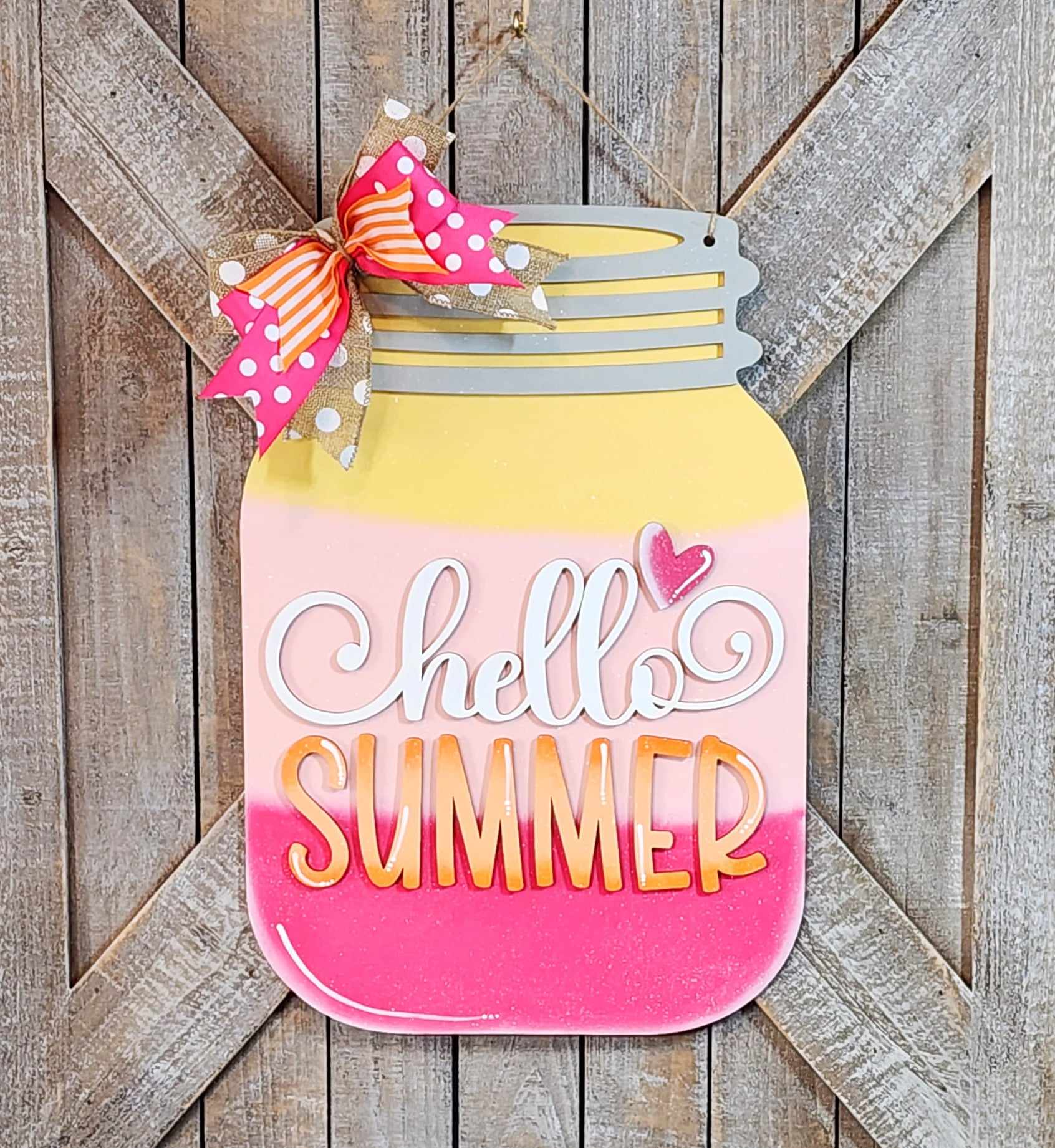 This delightful Hello Summer Mason Jar Door Hanger from Janet's Craft Corner showcases elegant cursive and bold lettering on a gradient backdrop of yellow, pink, and white. Accented with a polka-dotted ribbon bow at the top left, it is designed to hang on a rustic wooden surface with an X pattern—ideal for home décor or as part of a DIY Kit project.