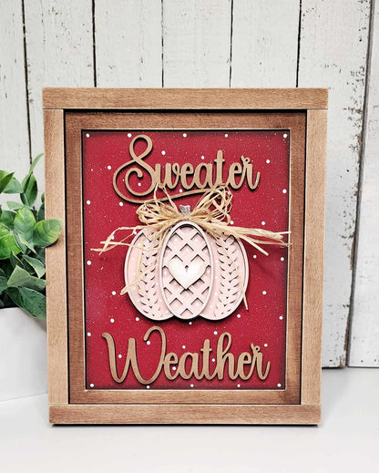 Introducing the "Sweater Weather Interchangeable Sign" by Janet's Craft Corner, a DIY home decor craft kit. This wooden-framed sign features "Sweater Weather" in elegant cursive on a vibrant red background, adorned with a decorative pumpkin topped with a bow at the center. Enhance your space effortlessly with this versatile decor piece, complementing any setting alongside a small potted plant against a white wooden wall backdrop.