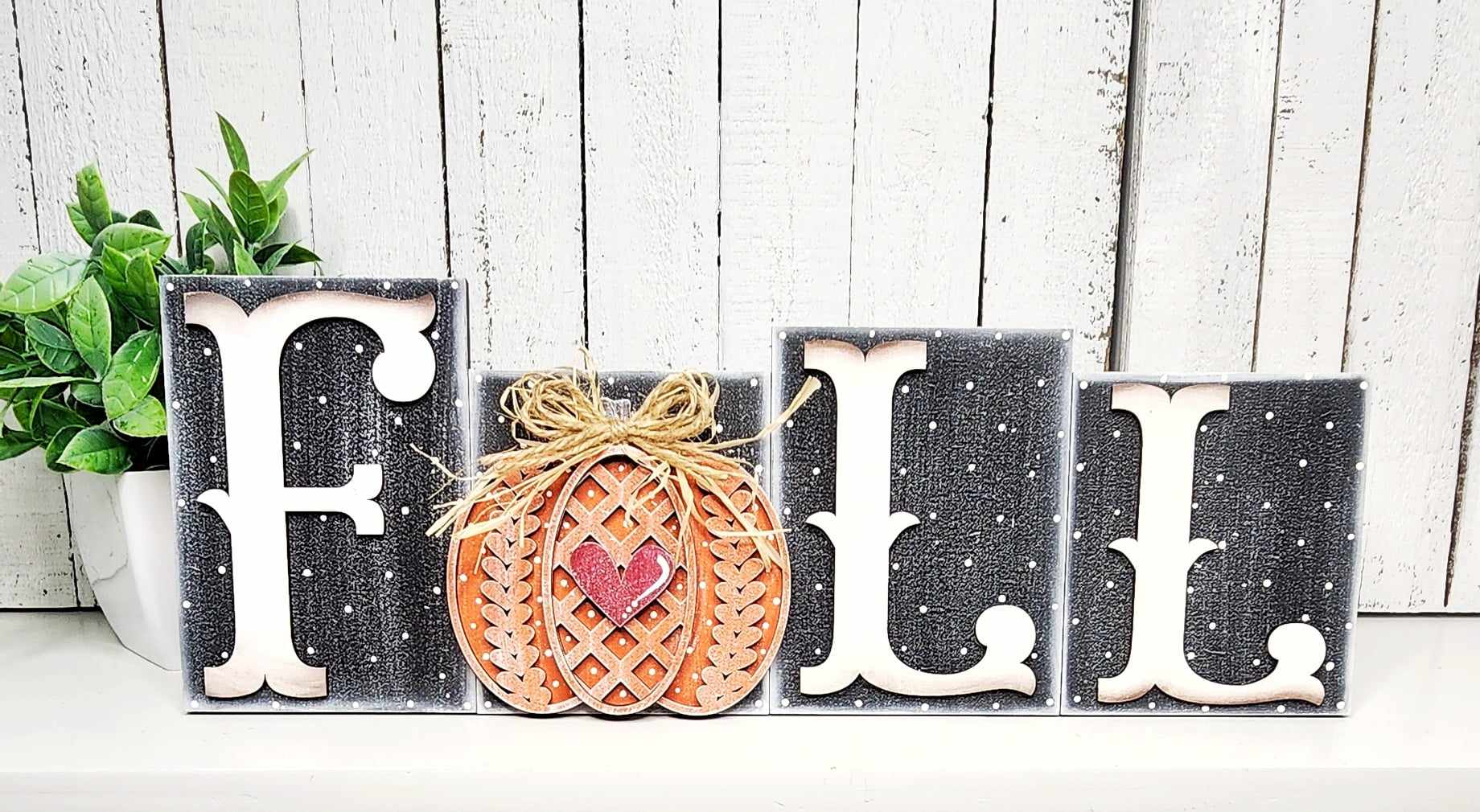 Janet's Craft Corner introduces the Fall Sweater Pumpkin Word Block DIY home decor craft kit, perfect for capturing autumn's charm. Against a rustic white wood backdrop, blocks spell "FALL," with the second block featuring a hand-painted pumpkin adorned with a red heart and twine bow. A small green plant on the left completes this handcrafted scene.