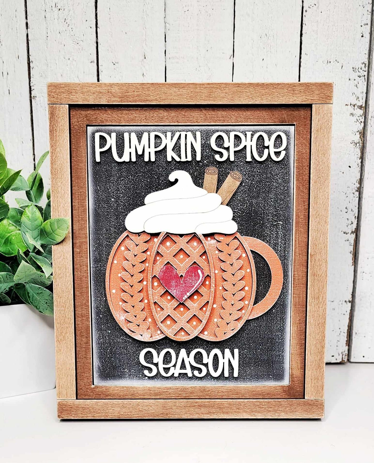 Immerse yourself in the cozy atmosphere with Janet's Craft Corner's Pumpkin Spice Season Interchangeable Sign. This DIY home decor craft kit features a decorative framed sign with a wooden mug, highlighted by a lattice pumpkin design, whipped cream, cinnamon sticks, and is adorned with a red heart. Embrace the spirit of "Pumpkin Spice Season" as text accompanies the design while a plant adds charm on the left.