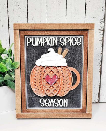 Immerse yourself in the cozy atmosphere with Janet's Craft Corner's Pumpkin Spice Season Interchangeable Sign. This DIY home decor craft kit features a decorative framed sign with a wooden mug, highlighted by a lattice pumpkin design, whipped cream, cinnamon sticks, and is adorned with a red heart. Embrace the spirit of "Pumpkin Spice Season" as text accompanies the design while a plant adds charm on the left.