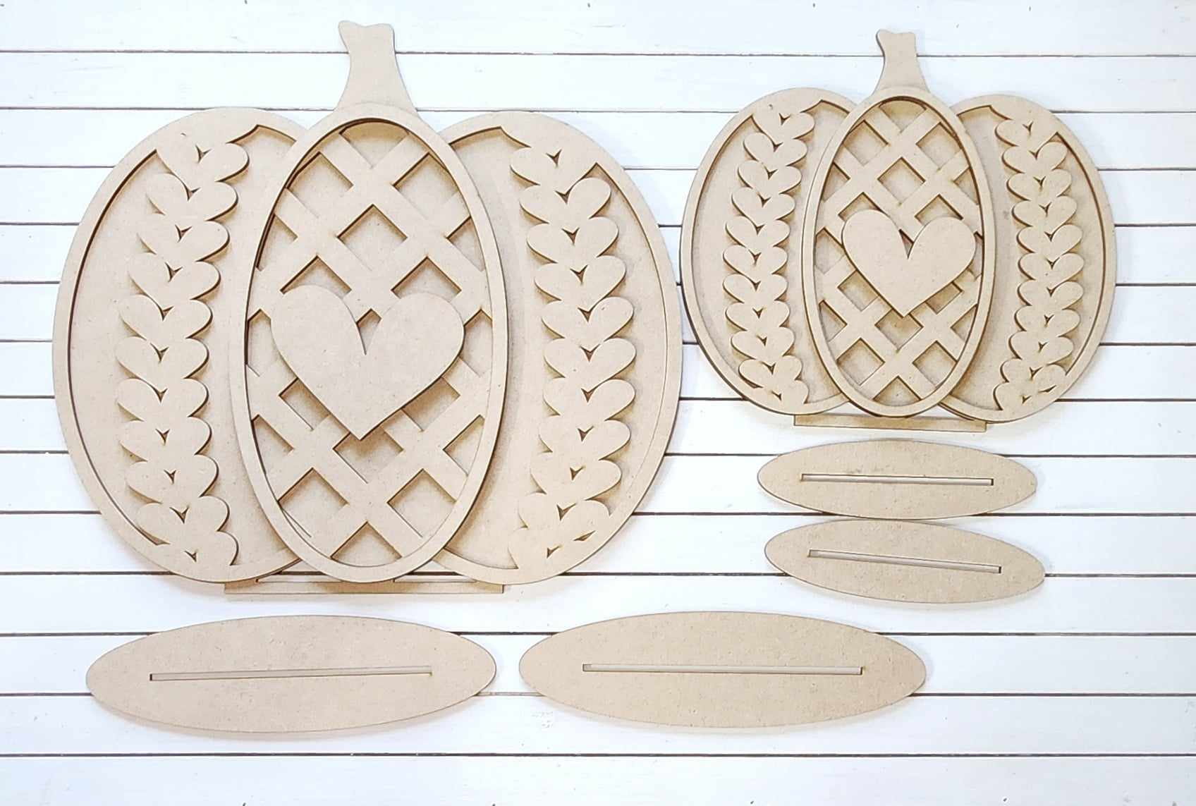 Janet's Craft Corner presents their Sweater Pumpkin Shelf Sitters, a set of three pumpkin-shaped wooden craft pieces with intricate lattice patterns and heart centers. Arranged on a white wood background, these delightful home décor accents come with detachable oval bases for easy assembly, making them the perfect DIY craft kit for your seasonal decoration needs.