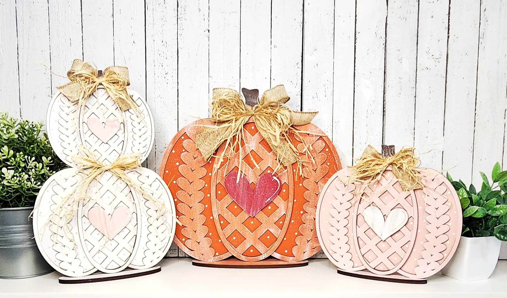 A set of three Sweater Pumpkin Shelf Sitters from Janet's Craft Corner showcases decorative pumpkins with intricate designs and bow accents against a white wooden background, ideal for enhancing any home décor. The pumpkins, displayed from left to right in white, orange, and light pink, are charmingly flanked by two green potted plants.