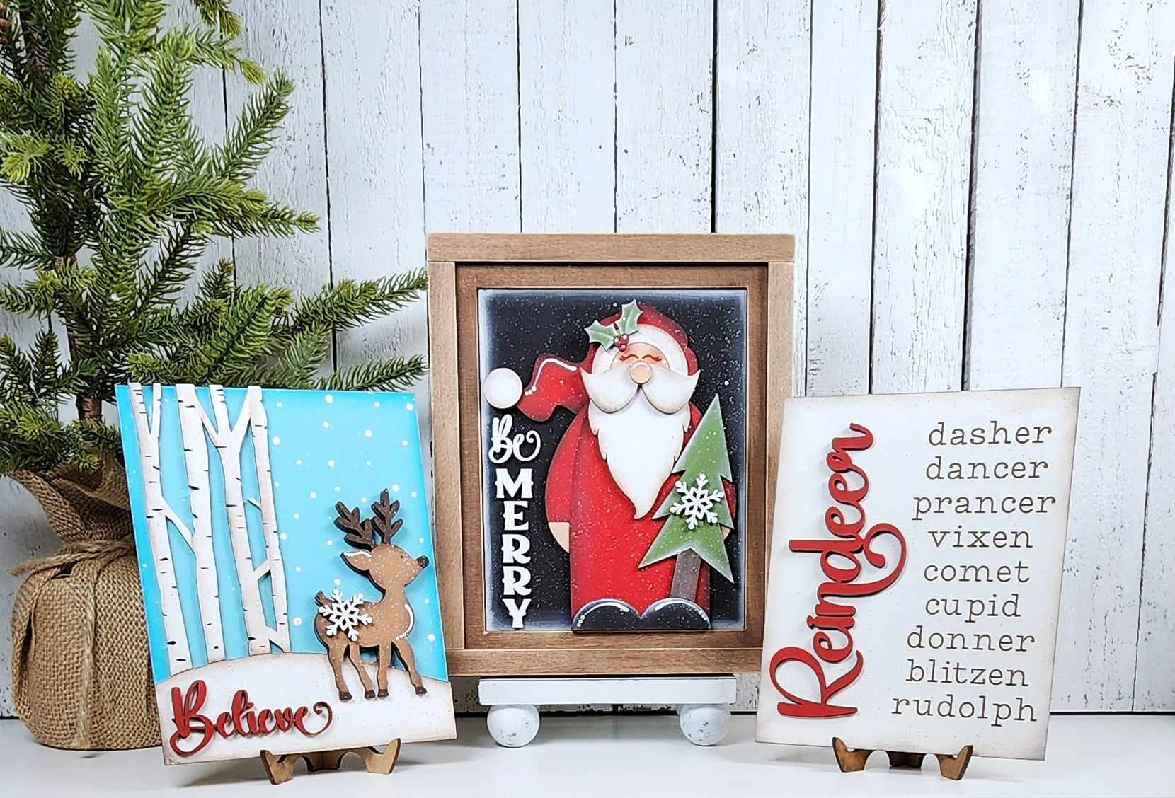 Janet's Craft Corner offers the Farmhouse Frame for Interchangeable Signs, a DIY home decor kit featuring a festive farmhouse style. It includes wooden plaques with designs: reindeer by birch trees with "Believe," Santa with "Be Merry," reindeer names with "Reindeer," and a small pine tree.