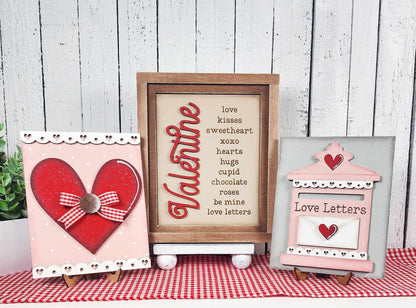 Three Valentine-themed decorations adorn the farmhouse-style table. Left: a red heart with a checked ribbon bow. Center: Janet's Craft Corner Farmhouse Frame for Interchangeable Signs featuring "love" and "kisses." Right: an envelope labeled "Love Letters." The background is a white wood wall.