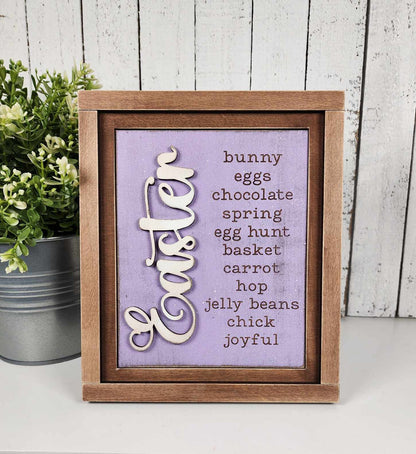 The Easter Words Farmhouse Sign by Janet's Craft Corner features a wooden frame with a purple background, showcasing words like "bunny," "eggs," and "joyful" alongside white cursive "Easter." Ideal for DIY decor, it includes a small potted plant and can enhance your craft kit.