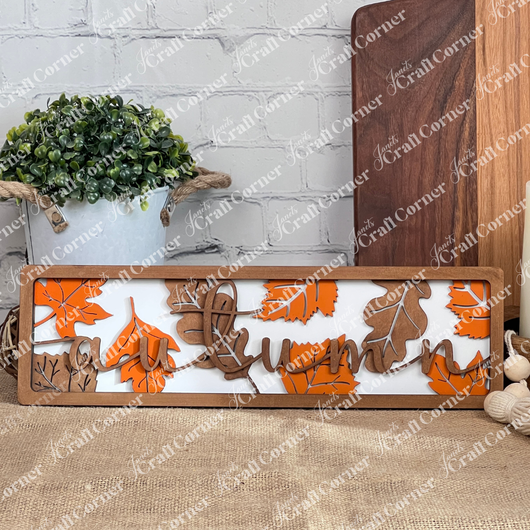 The Autumn Leaves Chunky Shelf Sitter by Janet's Craft Corner is a DIY home decor craft kit featuring wooden letters spelling "autumn," elegantly designed with orange and brown leaf accents. Perfect for adding a touch of seasonal charm, it complements potted plants and wooden cutting boards beautifully when displayed on a shelf.