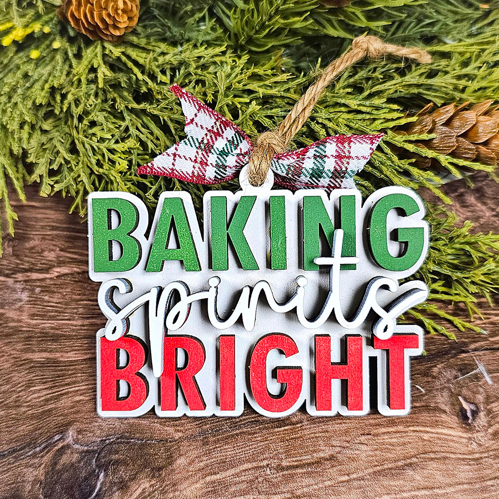 This individually painted ornament, named "Baking Spirits Bright" from Janet's Craft Corner, features the cheerful phrase in green, white, and red. Adorned with a plaid ribbon, it is beautifully displayed amidst evergreen branches and pinecones on a rustic wooden surface.