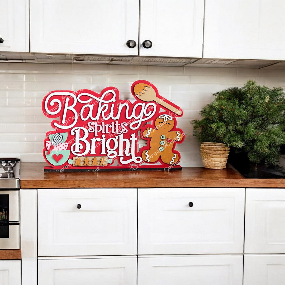 A festive kitchen countertop features Janet's Craft Corner "Baking Spirits Bright" shelf sitter with a gingerbread man and wooden spoon, alongside a potted evergreen plant. The white cabinets and subway tile backsplash enhance this DIY craft kit-inspired scene.