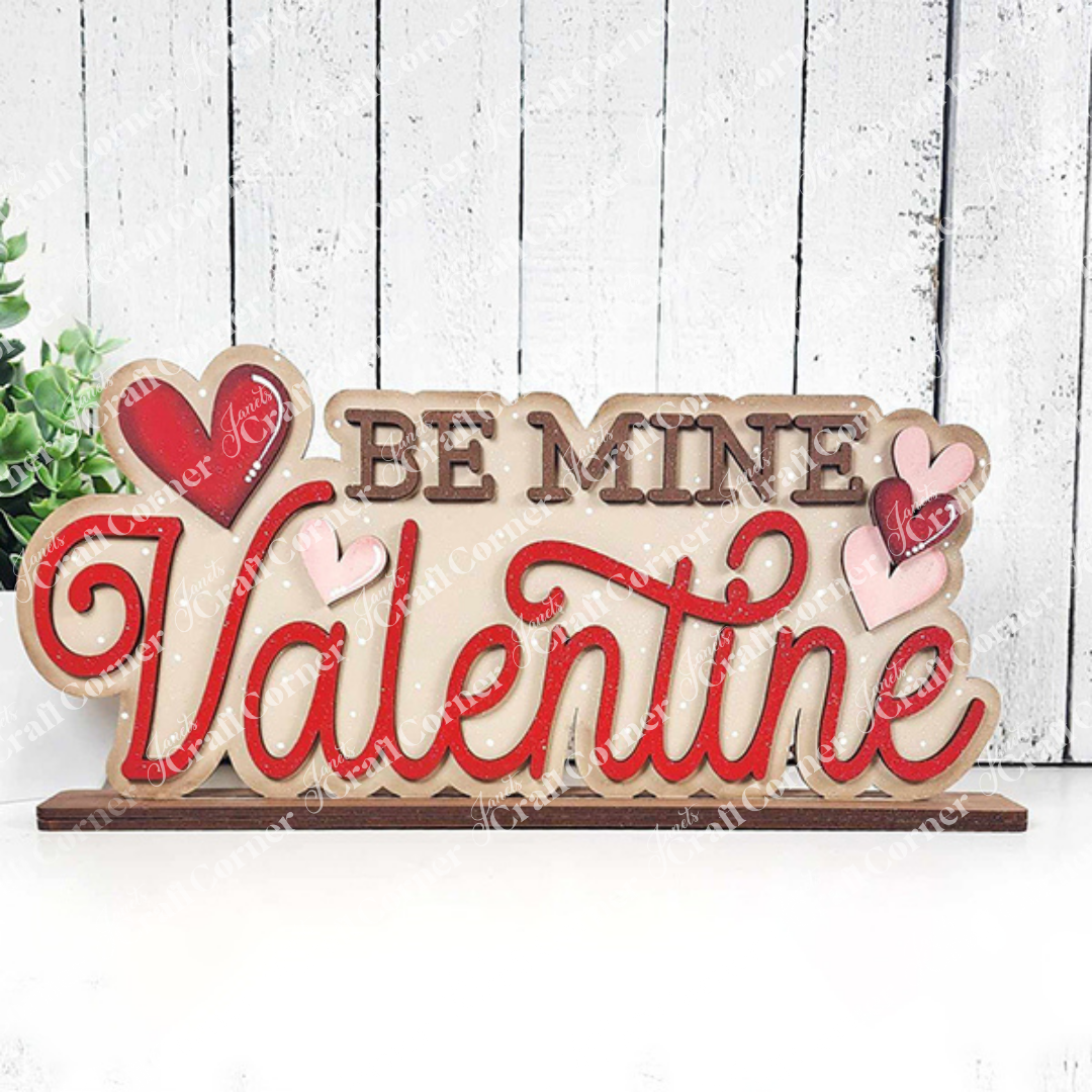 The "Be Mine Valentine Word Art" by Janet's Craft Corner features a hand-painted sign with "Be Mine Valentine" in red and brown, adorned with red and pink hearts. It stands charmingly against a rustic white wooden background, ideal for home décor.