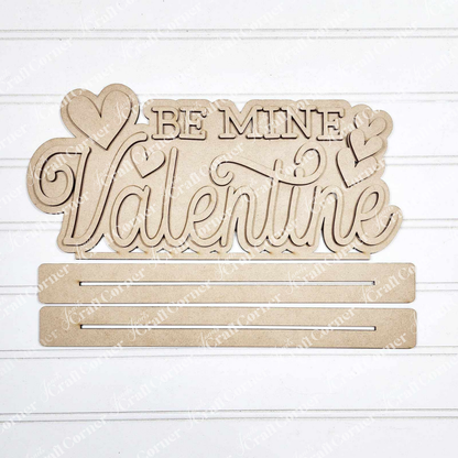 Add charm to your home décor with the "Be Mine Valentine" Word Art from Janet's Craft Corner. This hand-painted wooden cutout sign features cursive text and heart shapes on a white background, complete with two horizontal slots for DIY projects.