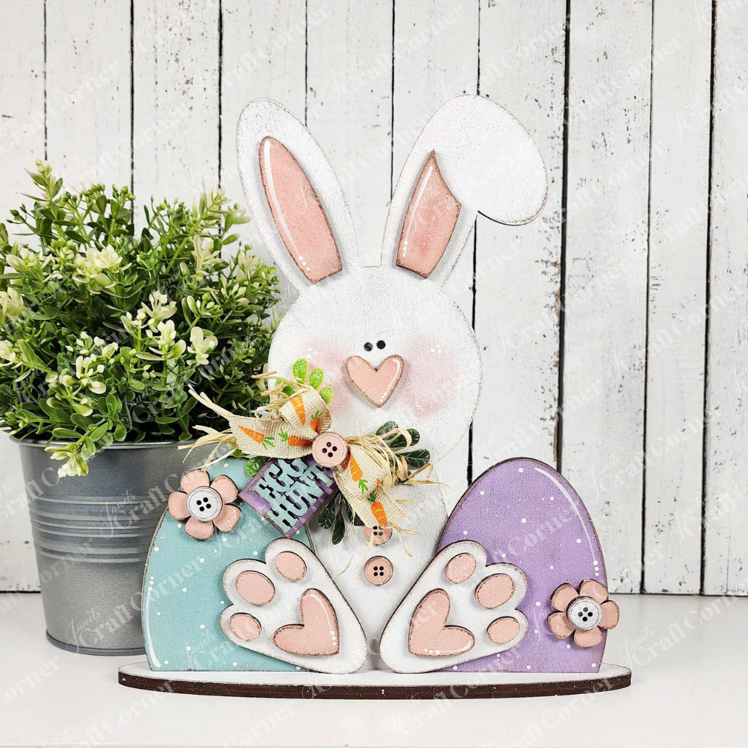 Janet's Craft Corner presents the Bunny Shelf Sitter, a DIY seasonal decor kit featuring a wooden Easter bunny with pastel pink and blue eggs, floral bow, and button details. It stands against a white panel with a potted plant for an enchanting finish.