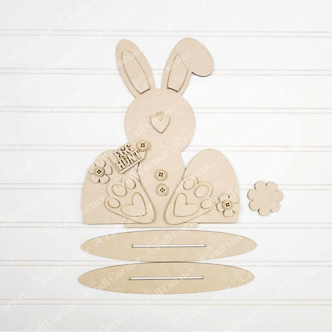 A flat-lay image displays Janet's Craft Corner's "Bunny Shelf Sitter" DIY craft kit on a white slatted backdrop, featuring wooden cut-outs: a bunny with buttons, a "Too Much Hunny" sign, a flower, and two base pieces.