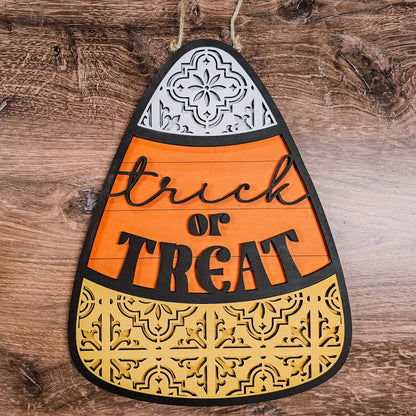 The Ornate Candy Corn Door Hanger from Janet's Craft Corner is a DIY home decor craft kit featuring the phrase "trick or TREAT" in script and block letters against a wooden background. This decorative piece includes three sections—white, orange, and yellow—adorned with intricate cut-out patterns.