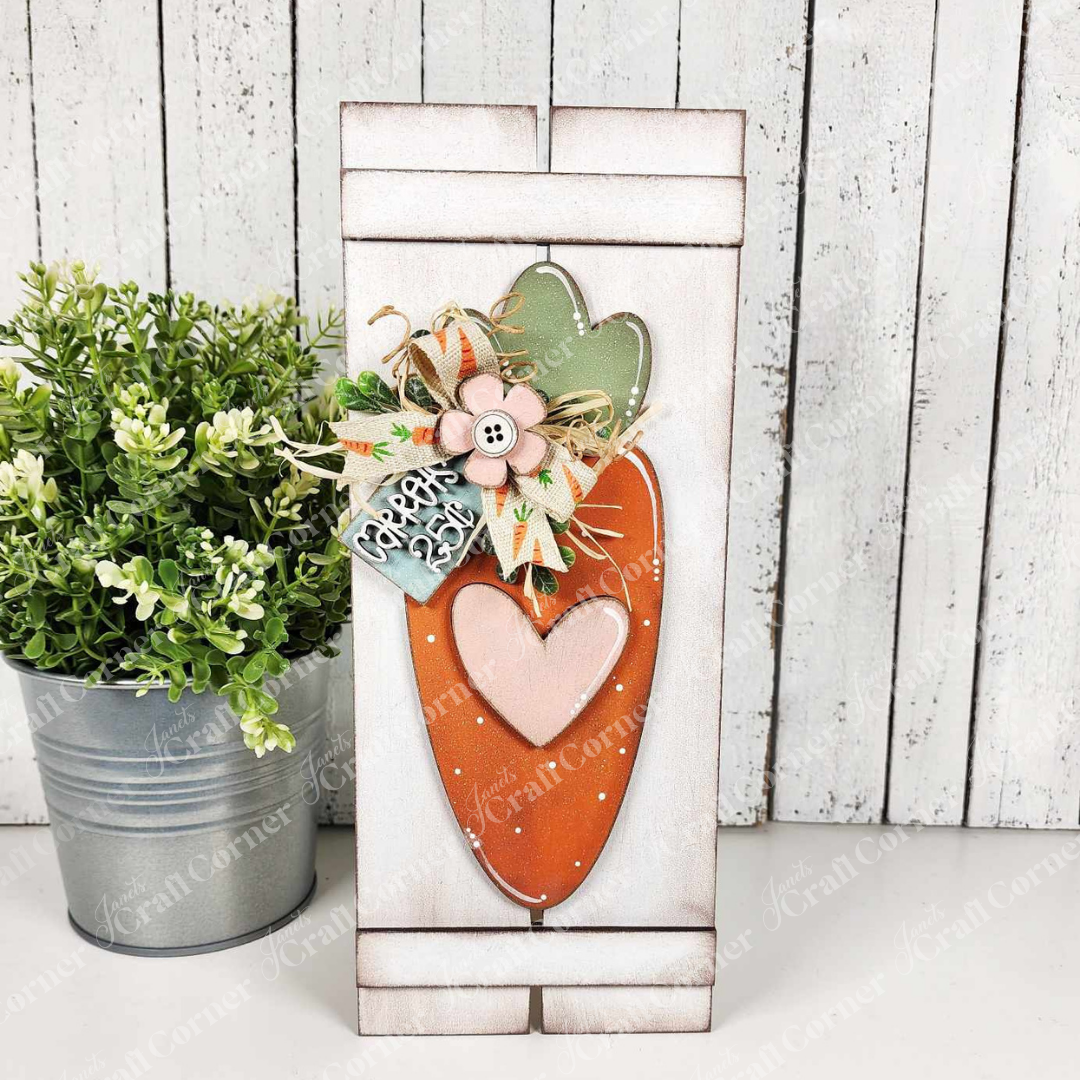 Janet's Craft Corner offers a charming Carrot Table Top Pallet sign with a painted carrot motif, hearts, and flowers. Enhanced by a floral bow and "cheerio" tag, it rests against a white wood backdrop beside a potted plant for seasonal decor.