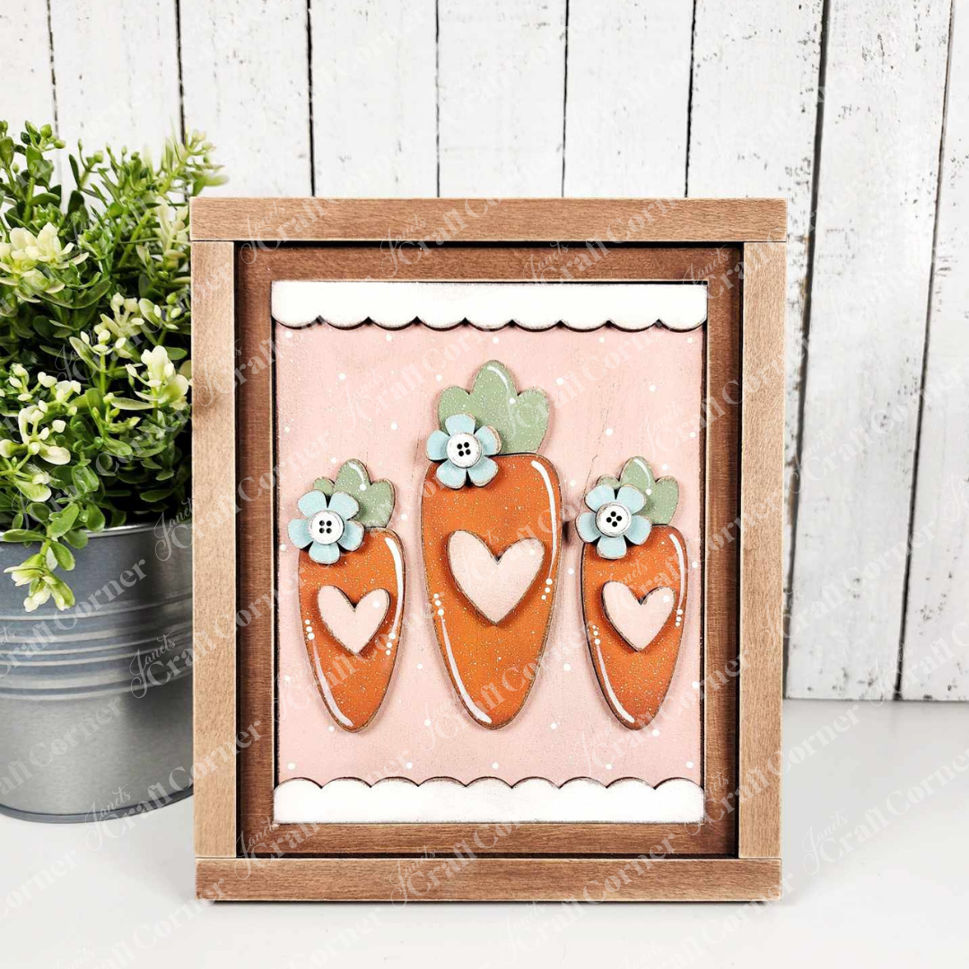 The Carrots Easter Farmhouse Sign by Janet's Craft Corner features three stylized carrots with heart cut-outs and blue flowers on a pastel pink background with white dots, complemented by a potted plant to the left and enclosed in a wooden frame.