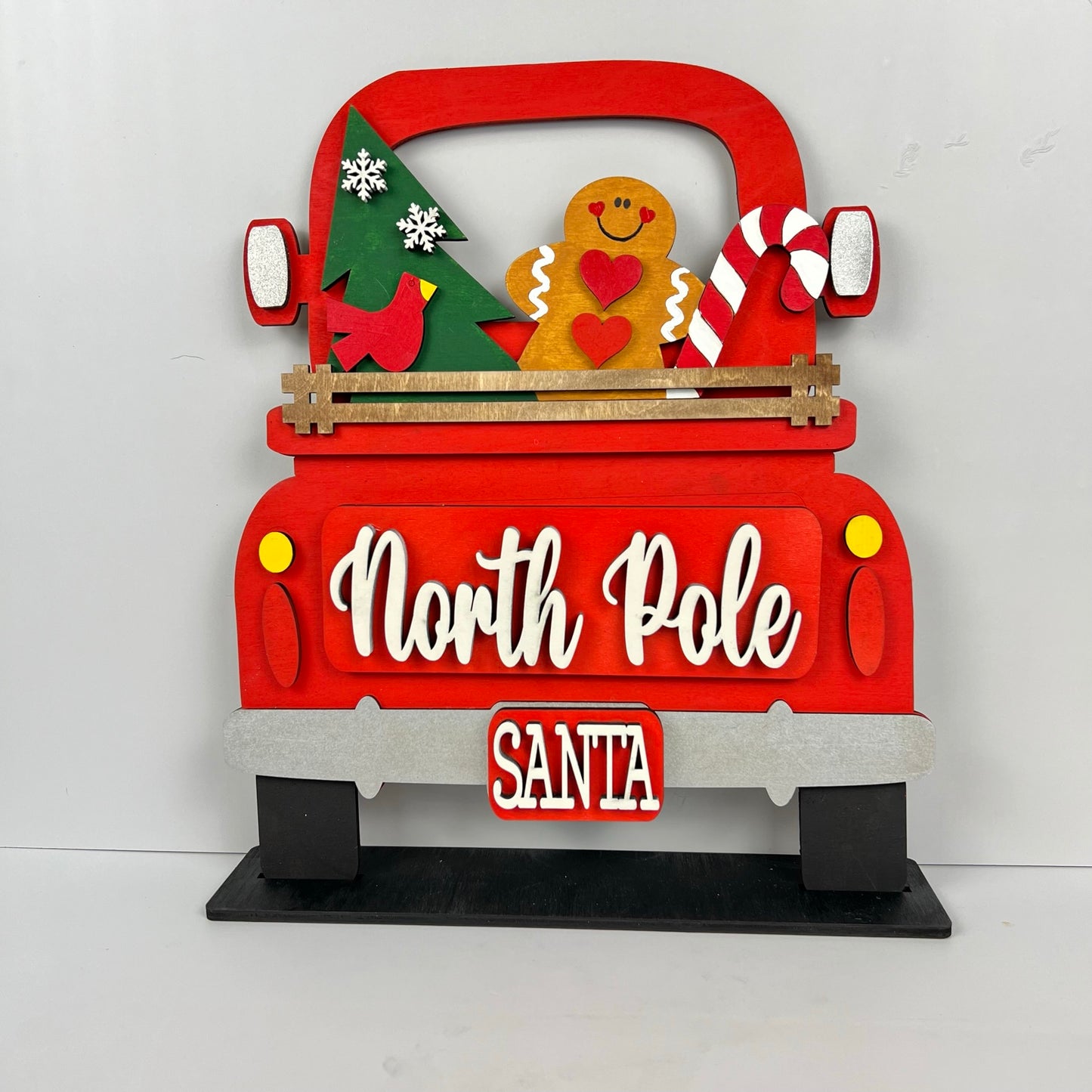 The "Antique Truck Base with Insert" by Janet's Craft Corner is a DIY craft kit featuring a festive red wooden truck cutout with a Christmas tree, gingerbread man, and candy cane. The tailgate reads "North Pole," and the license plate says "Santa." Ideal for interchangeable decor.