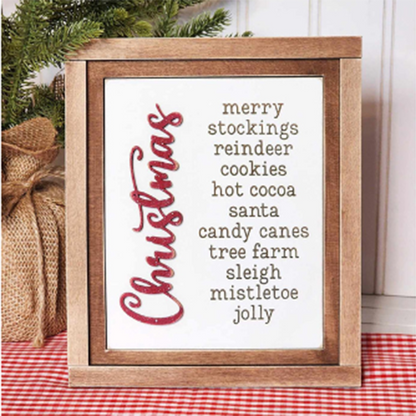 On a red checkered tablecloth, a sign from Janet's Craft Corner stands with a wooden frame, highlighting an interchangeable list of Christmas-themed words: merry, stockings, reindeer, cookies. Enhance your home decor with this DIY craft kit where "Christmas" is artistically styled vertically in red.