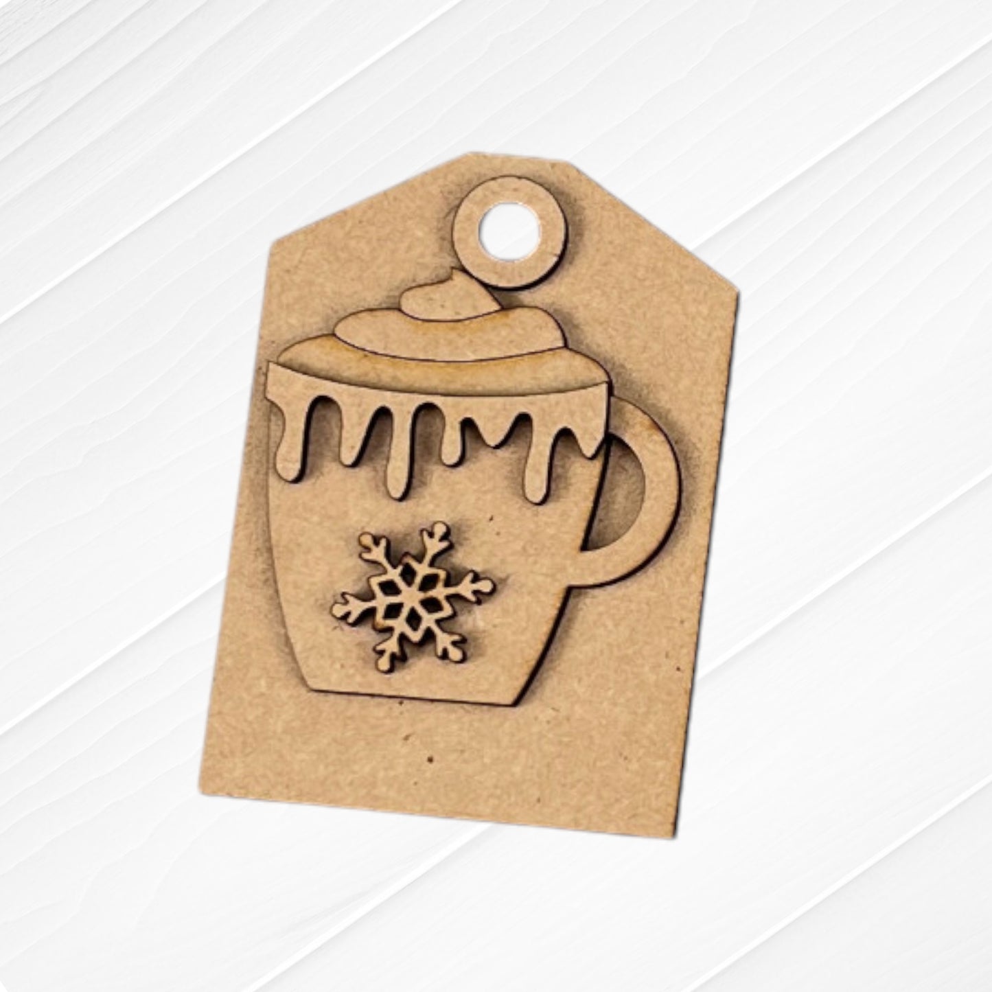 A cardboard cutout shaped like a gift tag highlights a mug with a snowflake design and frothy top, ideal for Janet's Craft Corner's Cup of Cheer Hot Cocoa Decor Set - Set of 5. The background features a white wooden surface, enhancing its cozy appeal.