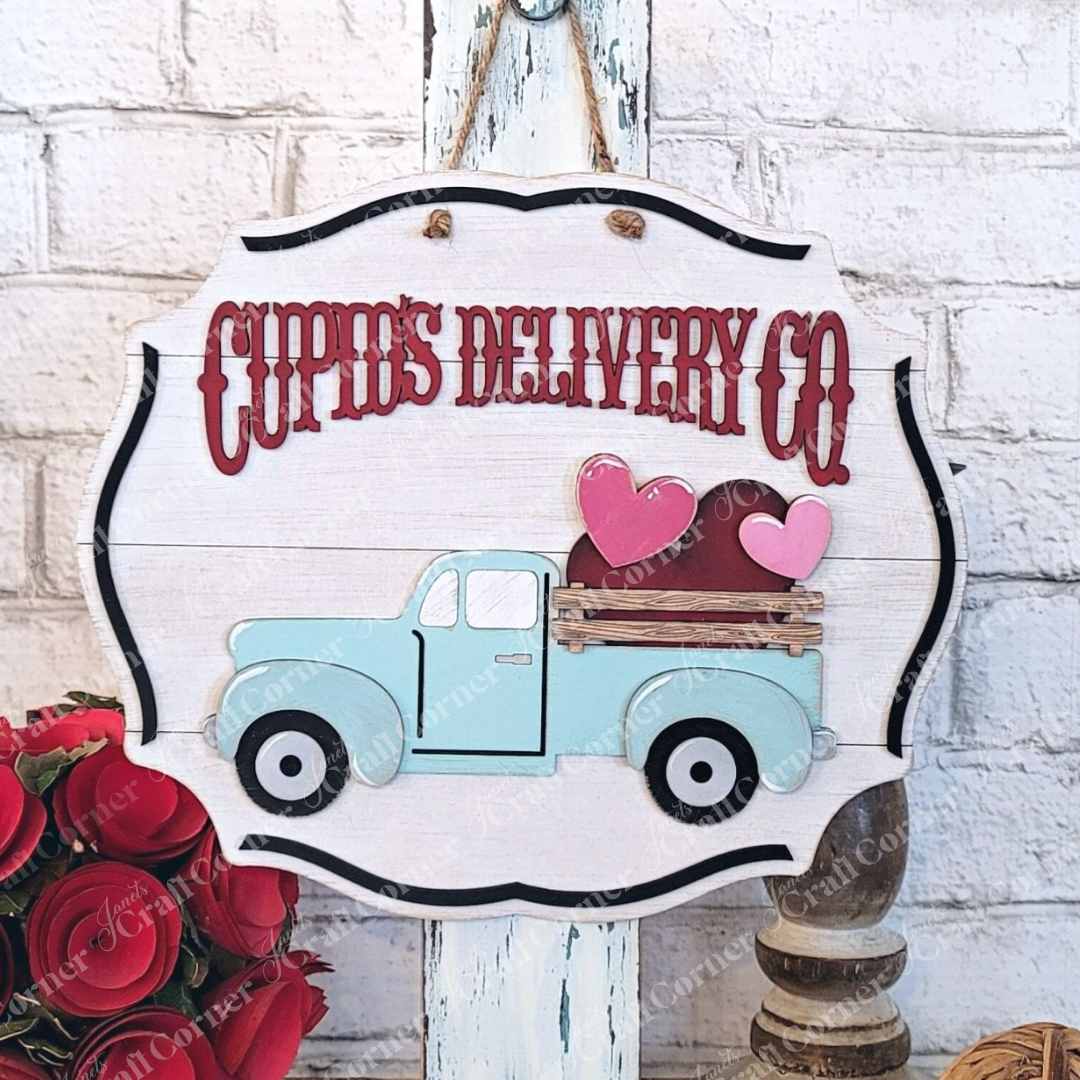 Introducing the Cupid Deliver Co from Janet's Craft Corner—a charming decorative wood sign designed especially for crafters. This vintage truck-shaped piece features "Cupid's Delivery Co" written above, with a light blue truck carrying hearts in the back. It's an ideal home décor item that beautifully complements a white brick wall.