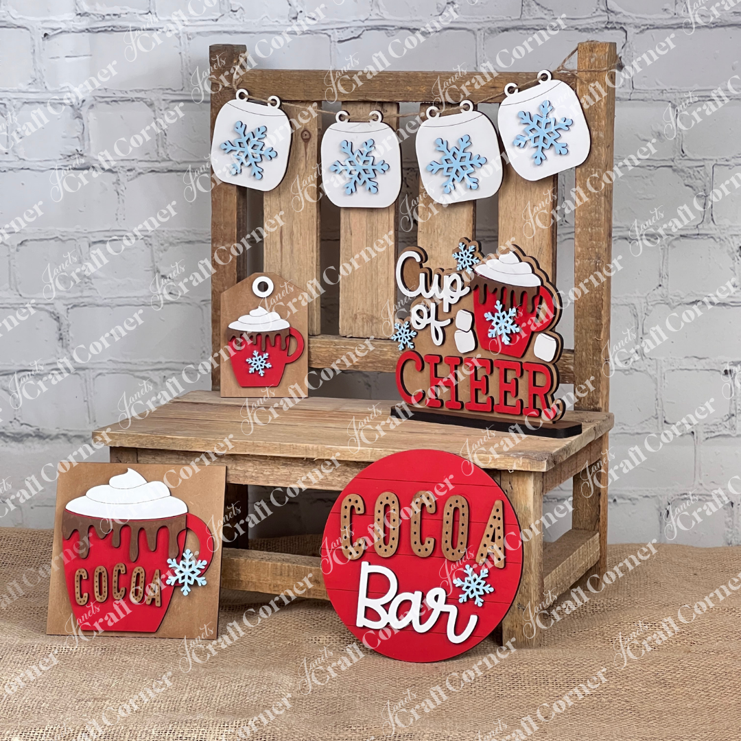 A festive wooden display from Janet's Craft Corner showcases the "Cup of Cheer Hot Cocoa Decor Set," featuring a hand-painted sign that reads "Cup of Cheer," accompanied by mugs of hot cocoa, snowflake embellishments, and a circular "Cocoa Bar" sign. Hanging above against the brick wall is a garland adorned with snowflakes, creating charming home décor from this DIY craft kit.