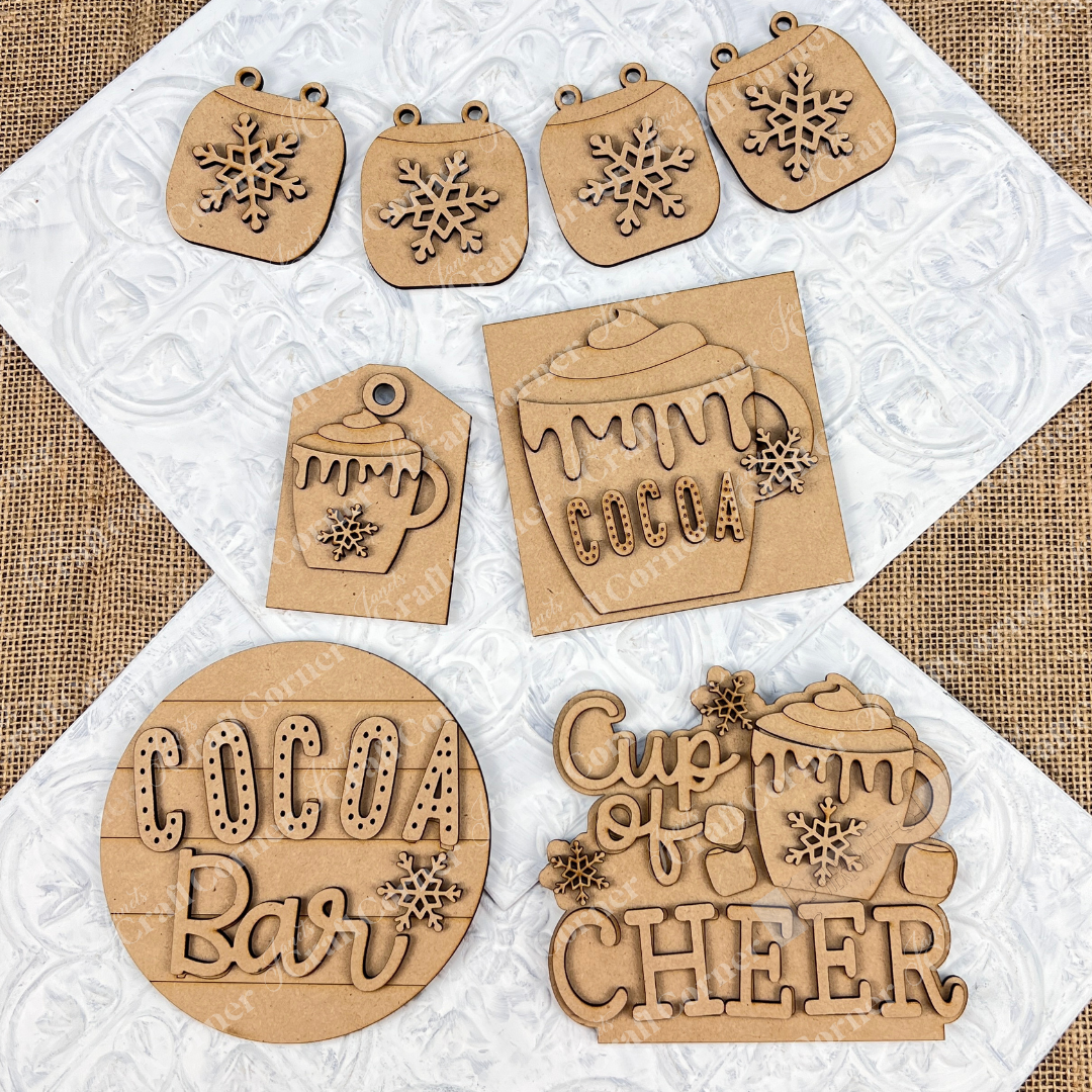 The Cup of Cheer Hot Cocoa Decor Set from Janet's Craft Corner is a DIY home decor craft kit featuring a collection of hand-painted wooden cutouts with holiday-themed designs, such as snowflakes, a "Cocoa" mug, and the phrases "Cocoa Bar" and "Cup of Cheer." Perfect for adding festive charm to your home, these pieces are beautifully displayed on a textured white surface.