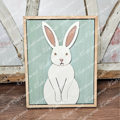 The Easter Bunny Farmhouse Sign by Janet's Craft Corner features a minimalist white rabbit with brown ears and eyes on a light green background. Its natural wood-like frame adds charm, making it perfect for seasonal decor against a rustic wooden surface.