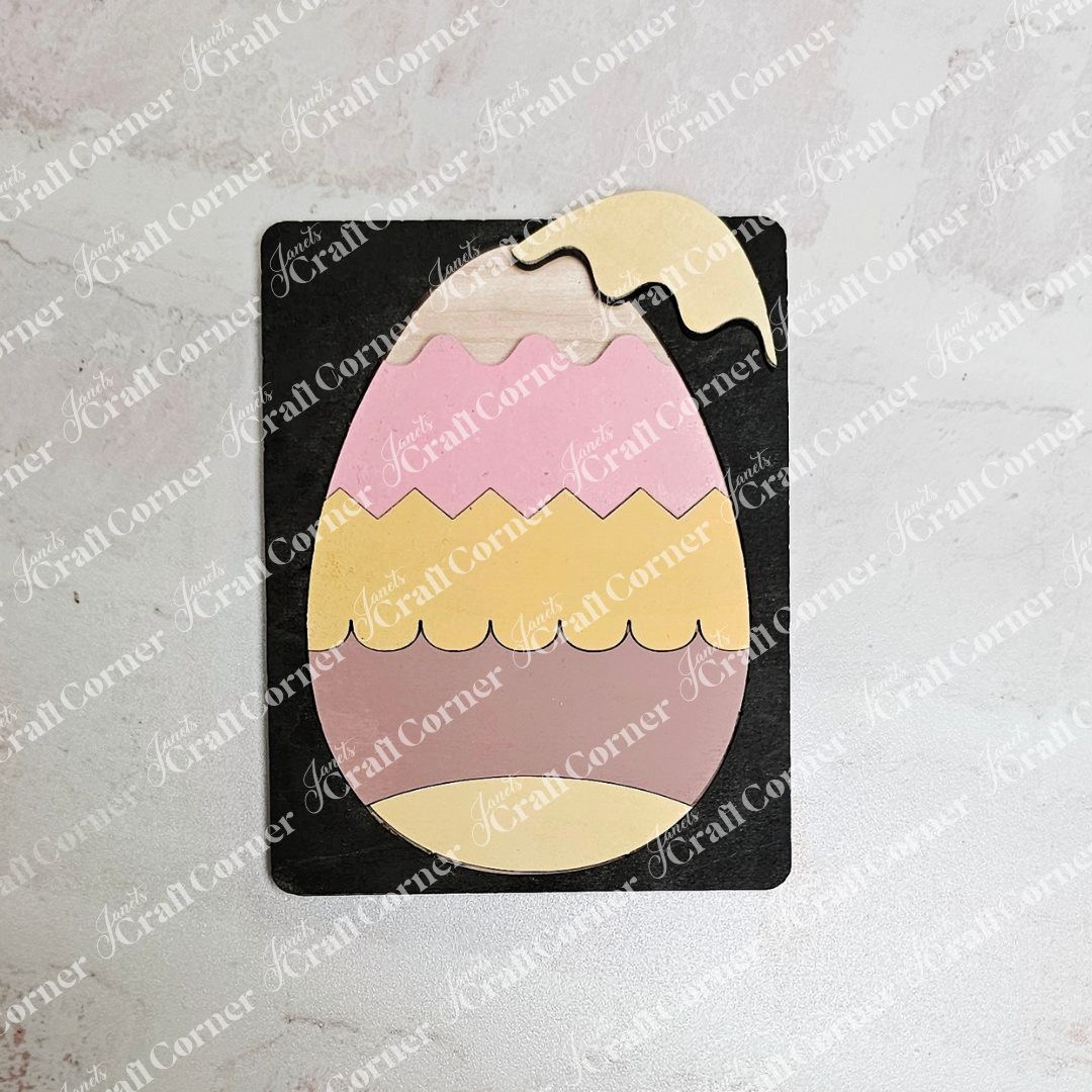 The Easter Egg Pop Out Puzzle from Janet's Craft Corner features a multicolored, egg-shaped design on a black background, resembling an Easter craft kit. The pastel cream, pink, and yellow layers are accentuated by a zigzag pattern. "KorantCorner" is watermarked across the image.