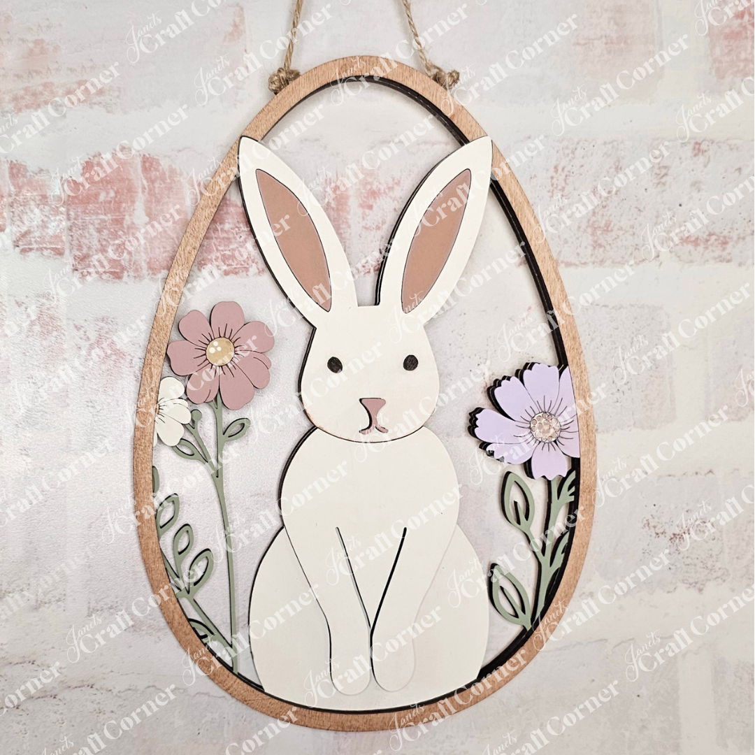 Janet's Craft Corner offers the Easter Egg Wall Hanger, a delightful wooden wall art featuring a white rabbit with long ears among pink and purple flowers on a light stone texture. Ideal for DIY seasonal home decor and crafting enthusiasts.