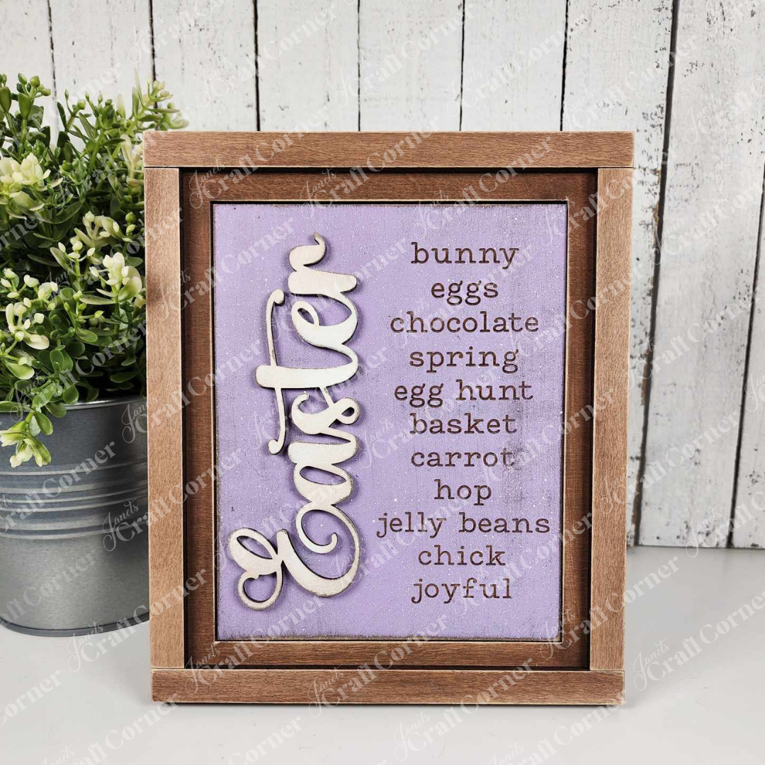 Janet's Craft Corner's Easter Switchable Farmhouse Signs Spring DIY Craft Kit features a purple sign with Easter words in black and "Easter" in large white script, displayed by a plant on a wood surface against a white wood panel background.