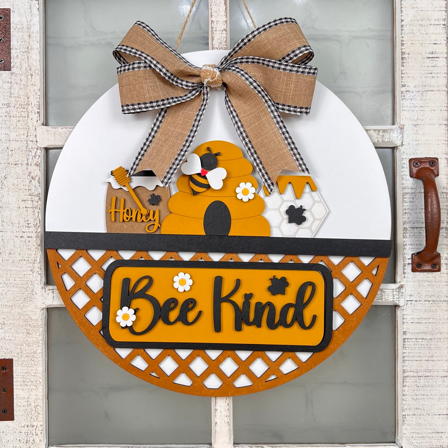 Janet's Craft Corner presents a DIY Door Hanger with a beehive design, bees, flowers, and "Honey Bee Kind" text. Includes checkered/burlap bow. Features hand-painted artistic decor on white lattice-like wood background. Set includes 1 interchangeable base and insert for creative home decor.