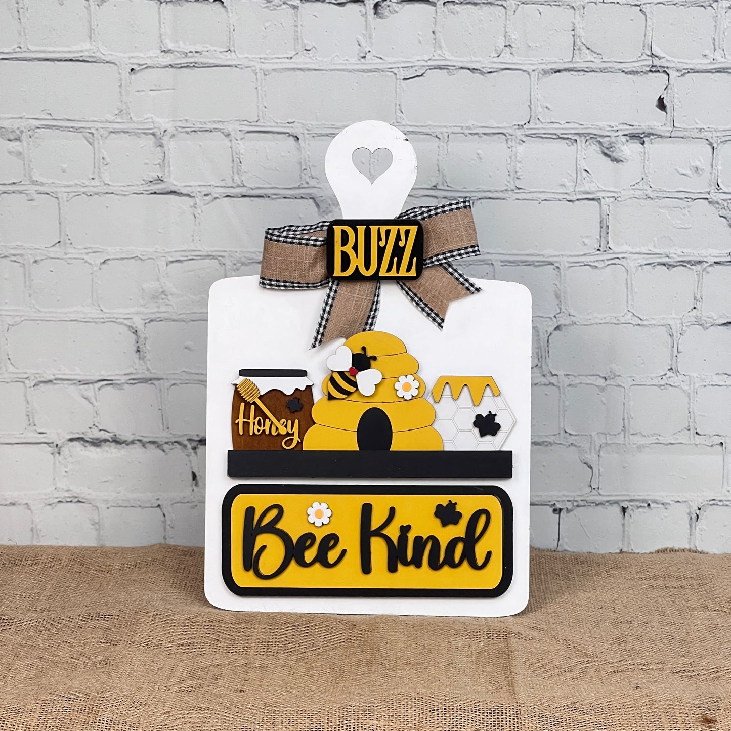 Enhance your home décor with Janet's Craft Corner DIY Breadboard Interchangeable Base with Insert featuring a bee theme. It displays a beehive, honey jars, and "Bee Kind" in yellow and black with a checkered "BUZZ" bow on a white brick wall background, perfect for your crafting needs.