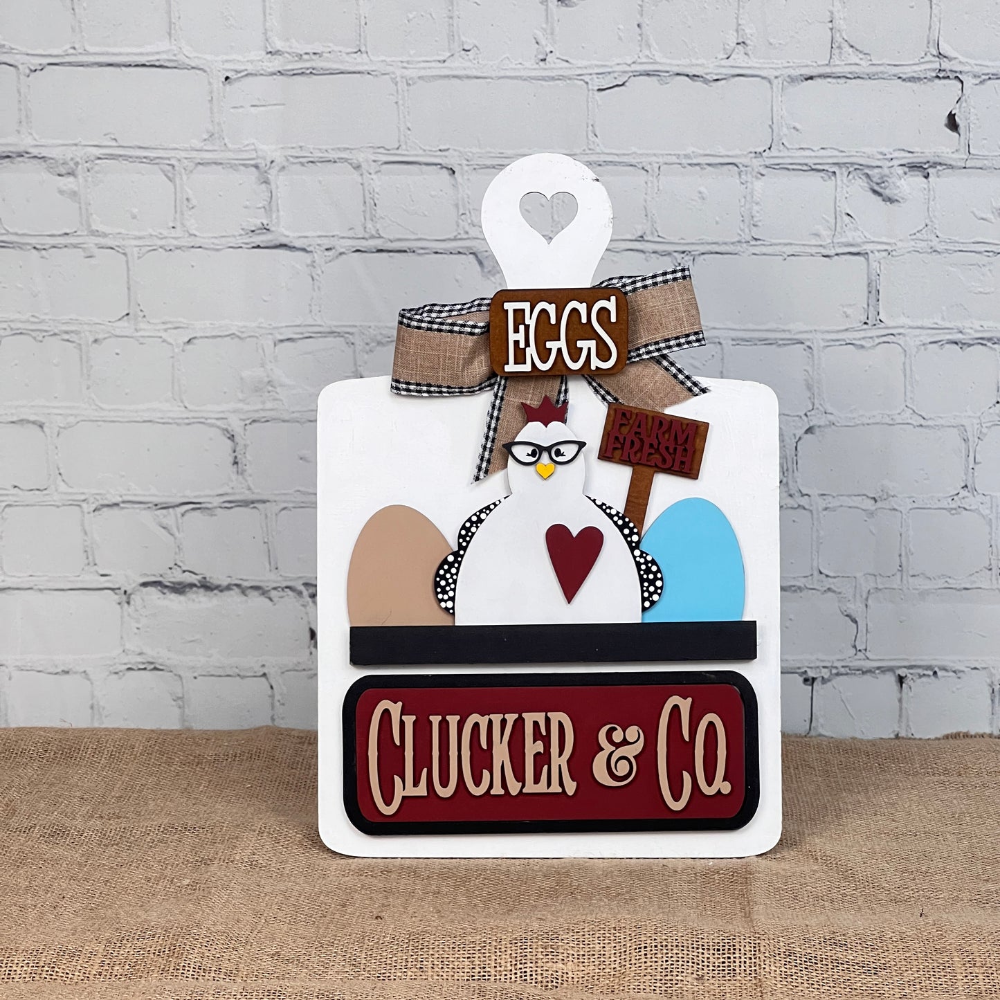 Janet's Craft Corner offers a charming DIY kit with a decorative breadboard featuring a cartoon chicken, eggs, and "Farm Fresh" sign. It reads "EGGS" on top and "CLUCKER & CO." below, with interchangeable heart and bow accessories against a rustic backdrop.