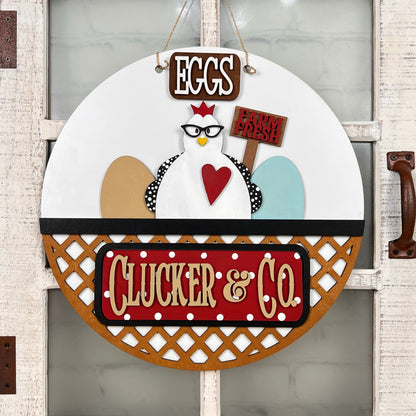 Janet's Craft Corner offers a DIY interchangeable door hanger featuring a cartoon chicken with glasses and a red crown holding "Farm Fresh" text. Above is "Eggs," and below, "Clucker & Co." appears in white on red with lattice detailing. Set includes 1 base and 1 insert.