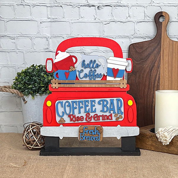 This charming home décor piece, by Janet's Craft Corner, features an antique truck base with a DIY interchangeable wooden red truck insert. Decorated with "Coffee Bar" and "Fresh Brewed" signs, coffee mugs, and hearts against a white brick wall, it's surrounded by cutting boards, a plant, and a candle.