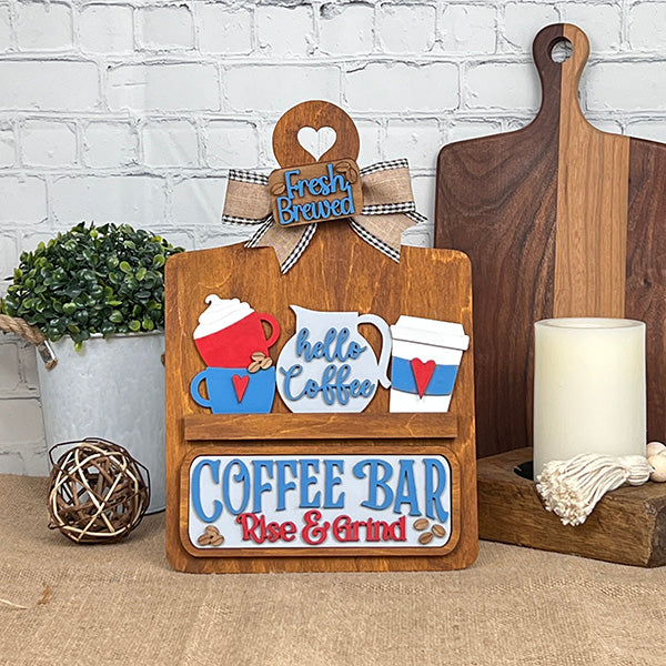 The Breadboard Interchangeable Base with Insert by Janet's Craft Corner features a decorative coffee-themed design with cutouts of coffee cups, a "Hello Coffee" pot, and "Coffee Bar, Rise & Grind" text. Comes adorned with a plaid bow, ideal for DIY décor enthusiasts seeking to enhance their home space.