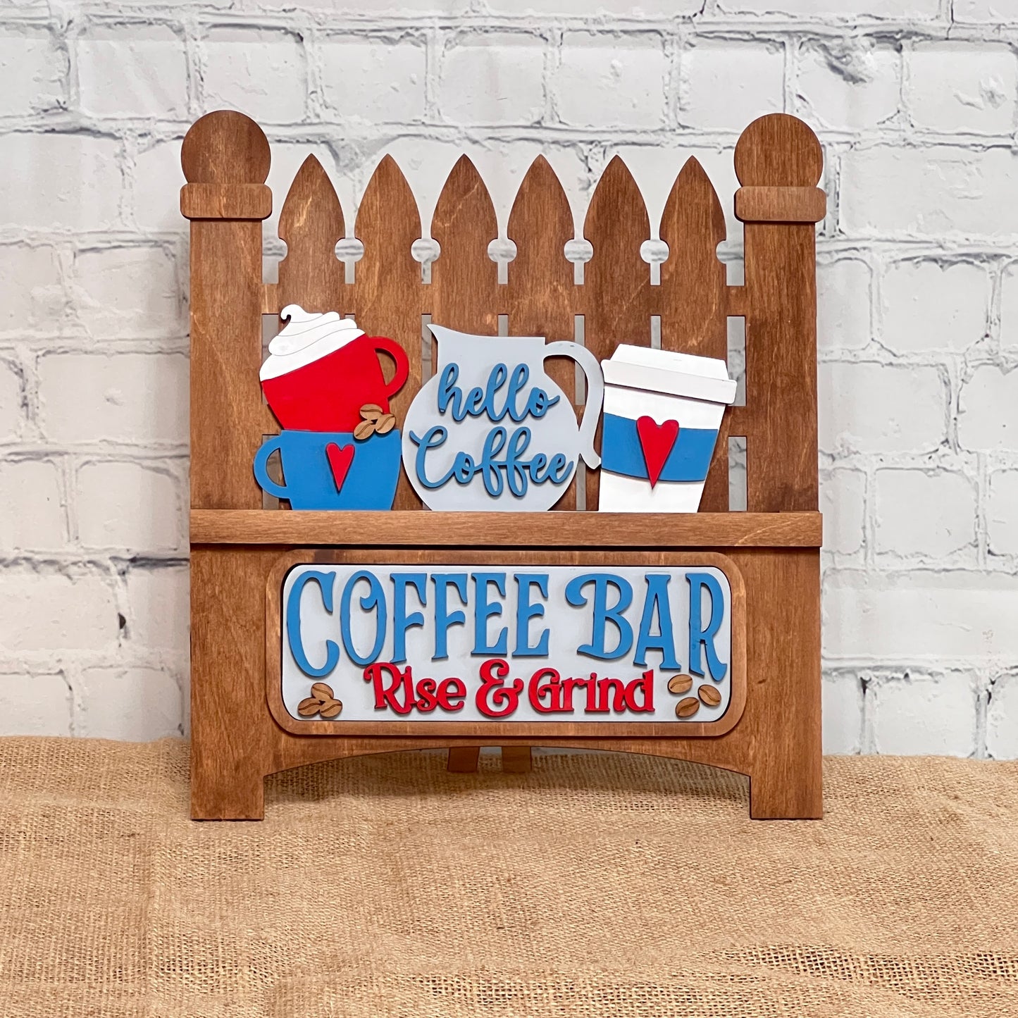 This delightful wooden sign from Janet's Craft Corner, resembling a classic picket fence, showcases coffee-themed designs with cups and a pot. It reads "Hello Coffee" and "COFFEE BAR Rise & Grind," set against a white brick background on burlap, ideal for DIY craft kit enthusiasts.