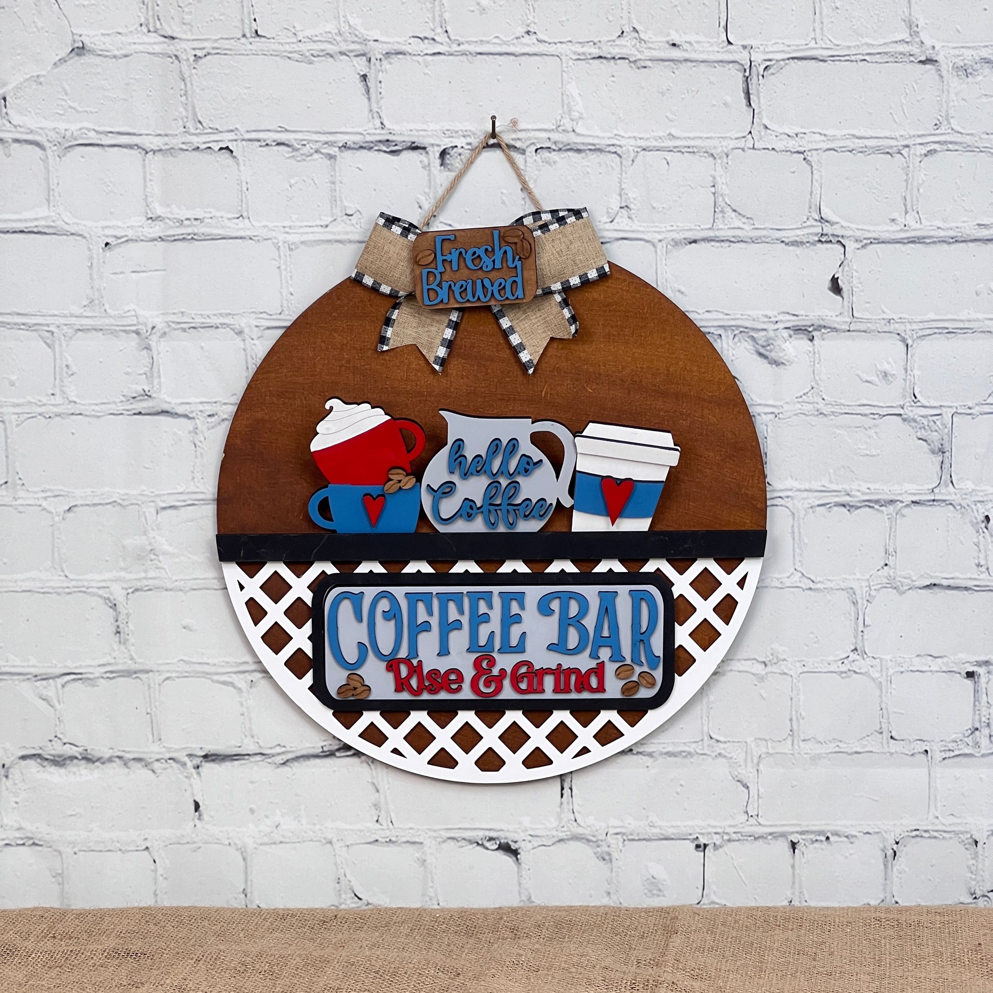 This delightful interchangeable door hanger from Janet's Craft Corner features a round wooden sign adorned with coffee-themed decorations, including hand-painted cups, a kettle, and phrases like "Fresh Brewed" and "COFFEE BAR, Rise & Grind," all topped with a bow.