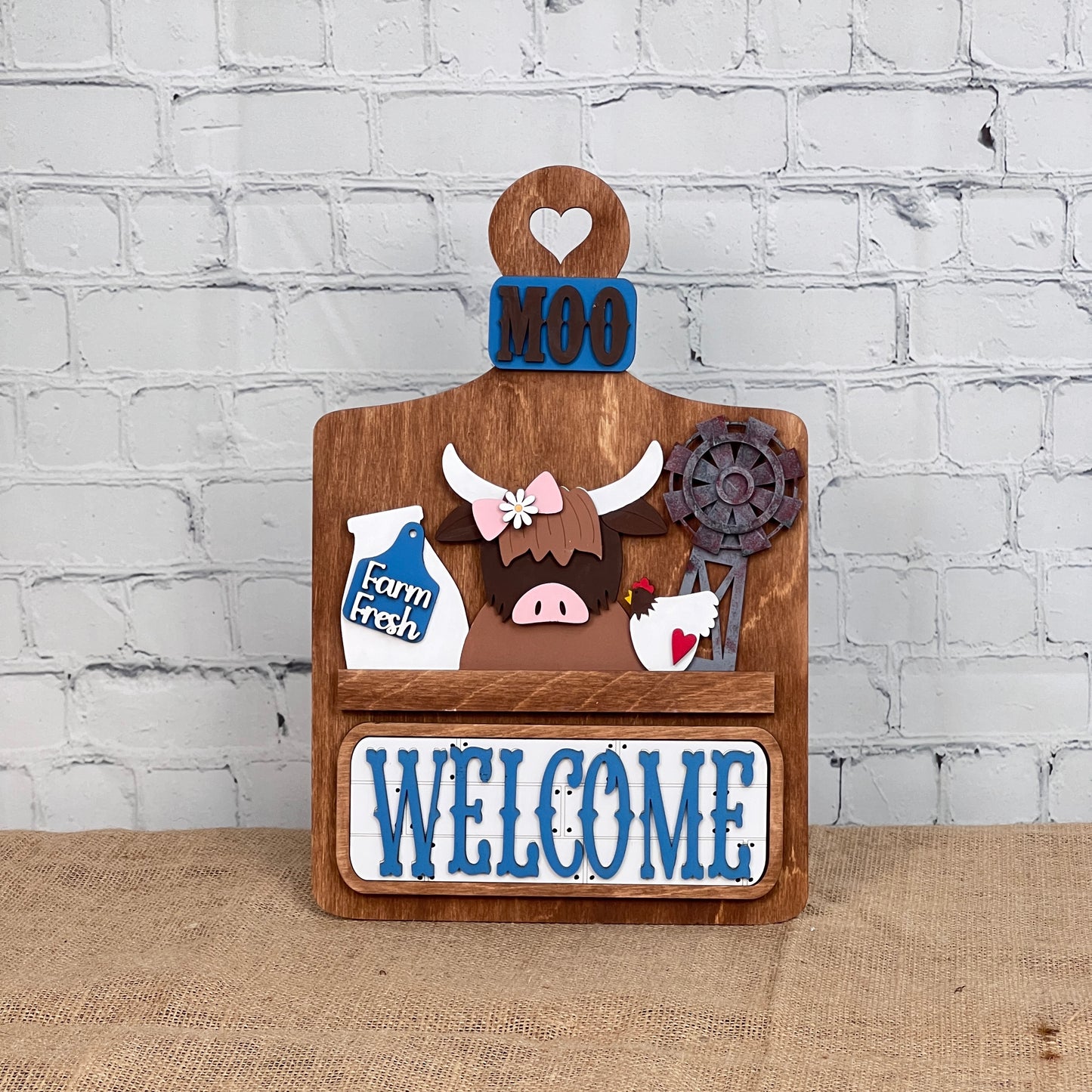 The Breadboard Interchangeable Base with Insert by Janet's Craft Corner is a wooden home décor kit featuring "MOO" and a cow graphic. It includes accessories like a "Farm Fresh" milk jug, windmill, and hen, with "WELCOME" displayed on burlap against white brick.