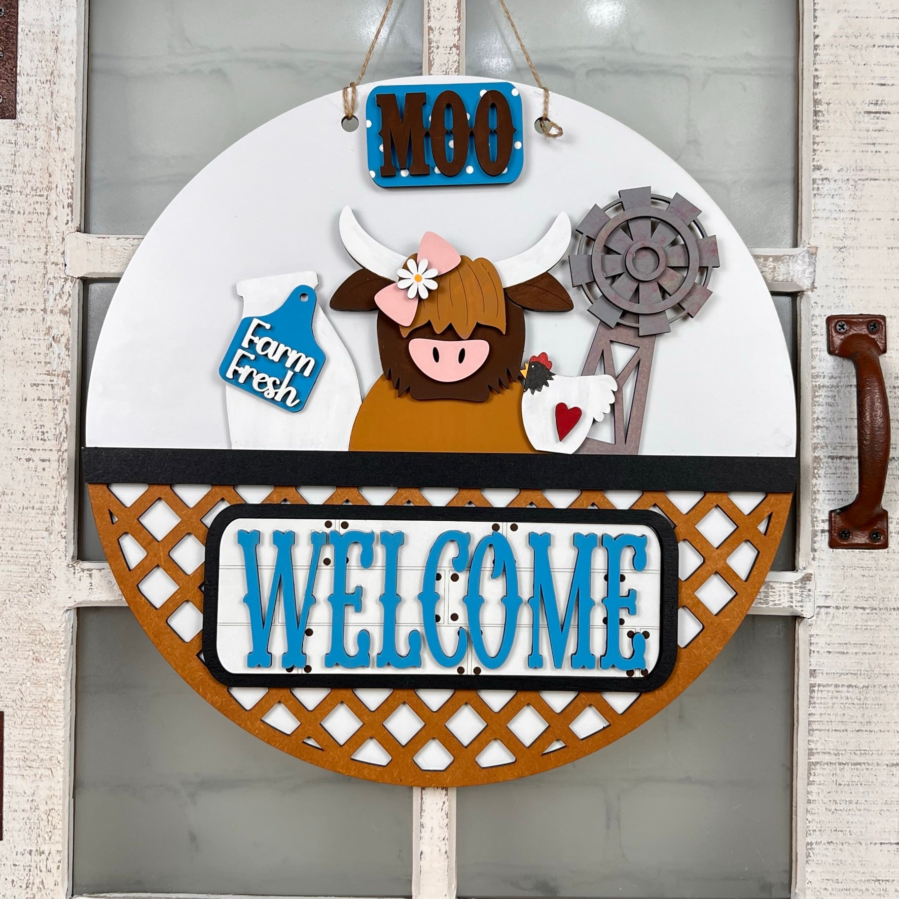 Janet's Craft Corner offers a round, decorative wooden sign perfect as an interchangeable door hanger. It has a cartoon cow with a cowbell, windmill, milk jug, and the words "MOO," "Farm Fresh," and "WELCOME" on a rustic whitewashed door. Set includes 1 door hanger base and 1 insert.