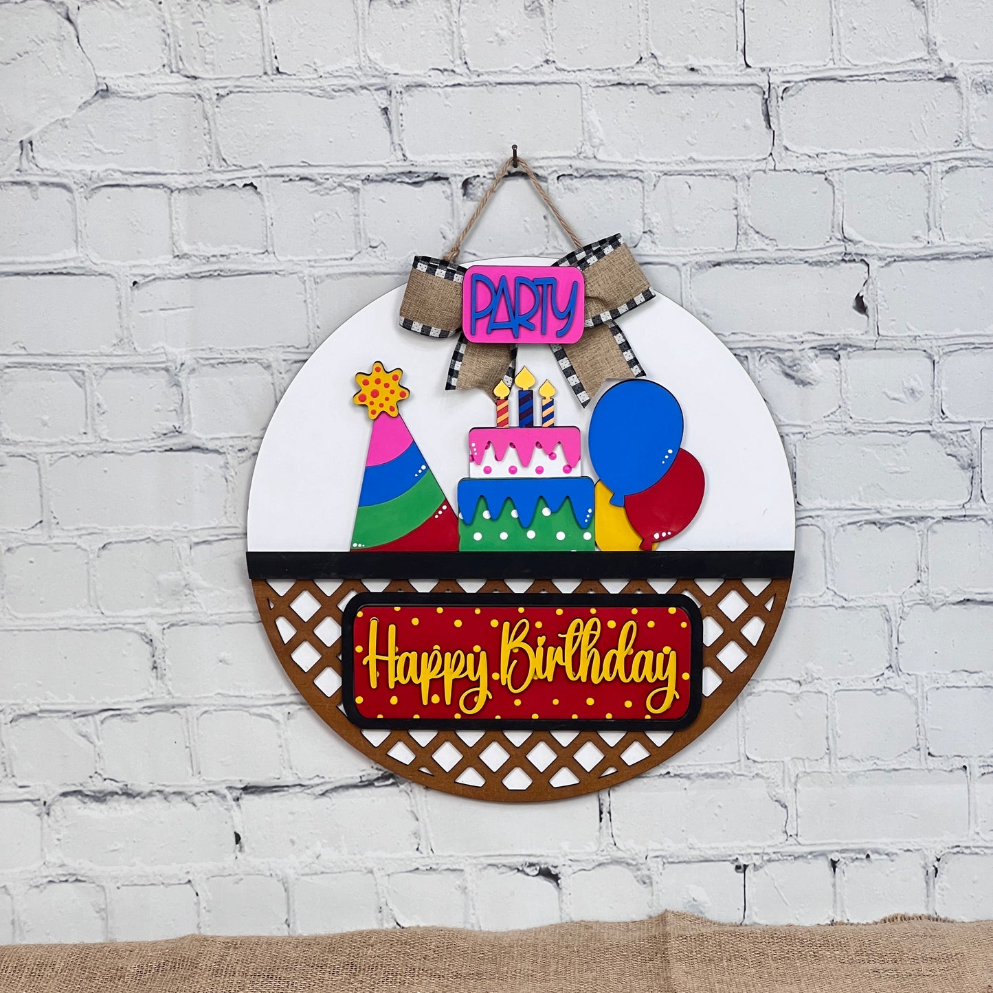 A Janet's Craft Corner DIY kit showcases a decorative birthday sign against a white brick wall. It features a colorful cake, candles, balloons, and party hat with "Party" on top and "Happy Birthday" below on red. Ideal for an interchangeable door hanger project!.