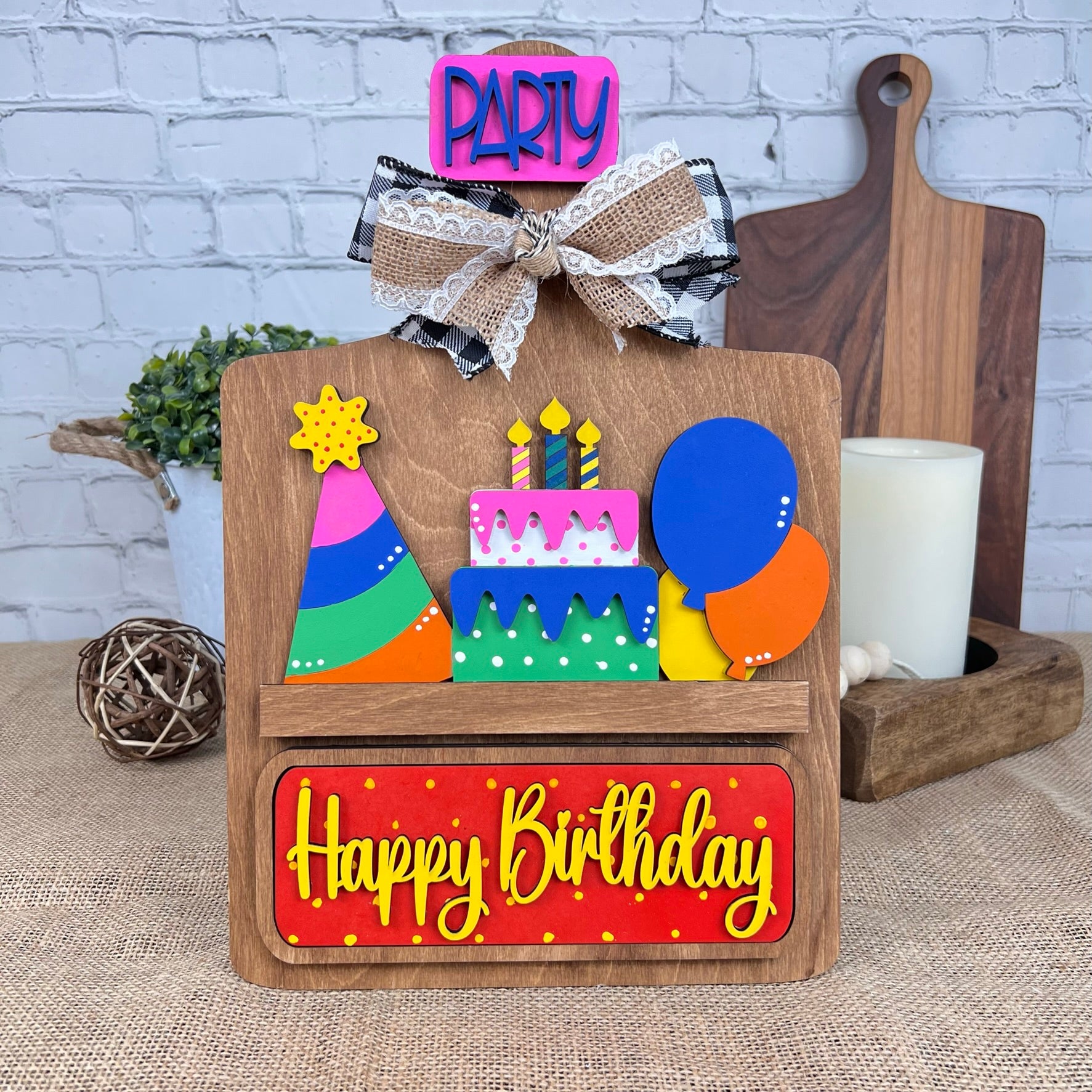 The Breadboard Interchangeable Base with Insert by Janet's Craft Corner is a DIY home decor kit featuring a wooden birthday theme with colorful elements like a cake and balloons, complete with "Happy Birthday" and "Party" signs, plus interchangeable accessories for personalized décor.