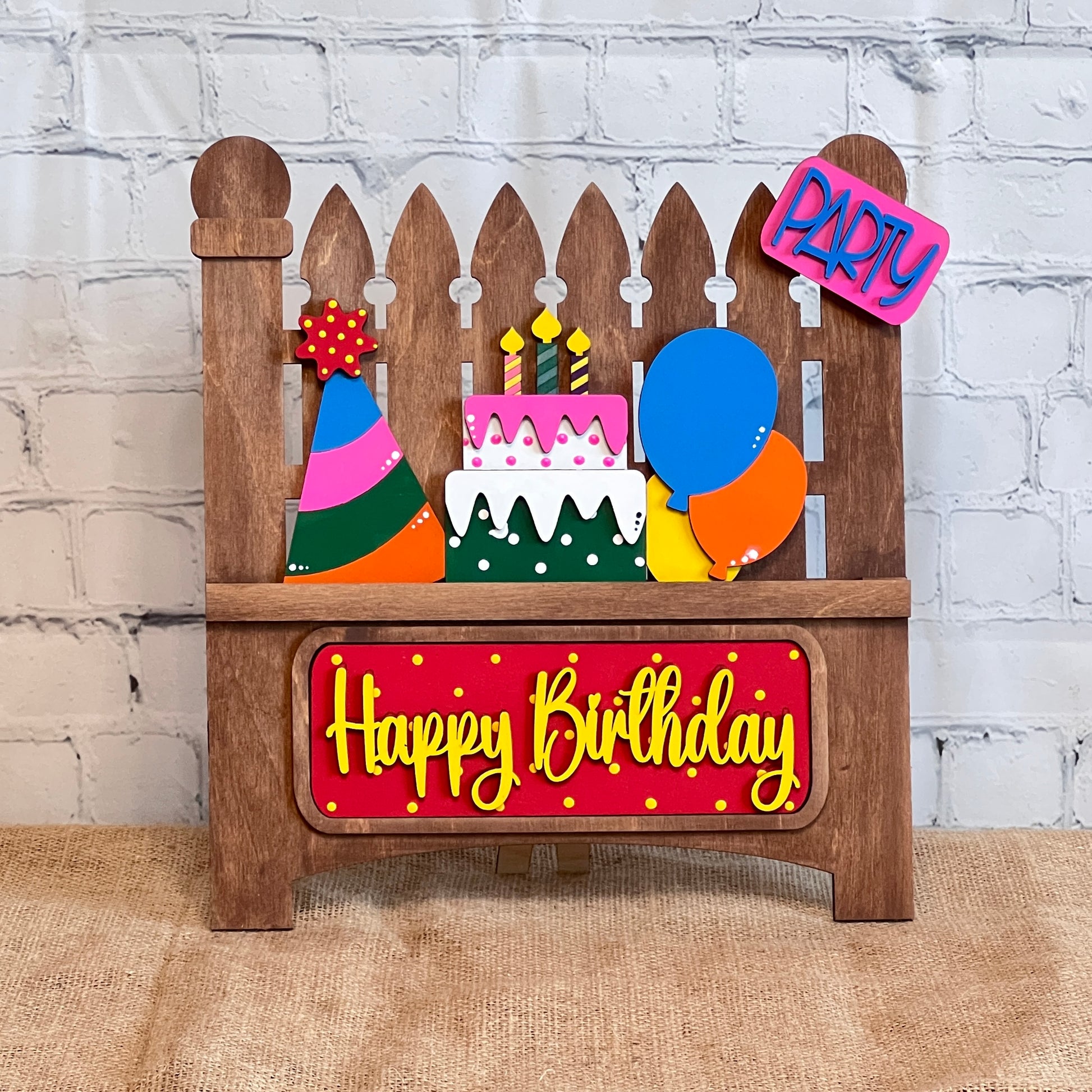 Janet's Craft Corner offers a DIY kit featuring a wooden birthday decoration with a picket fence backdrop. It includes a layered cake, vibrant balloons, festive hats, and signs that say "Party" and "Happy Birthday." Ideal for crafting lively ensembles.