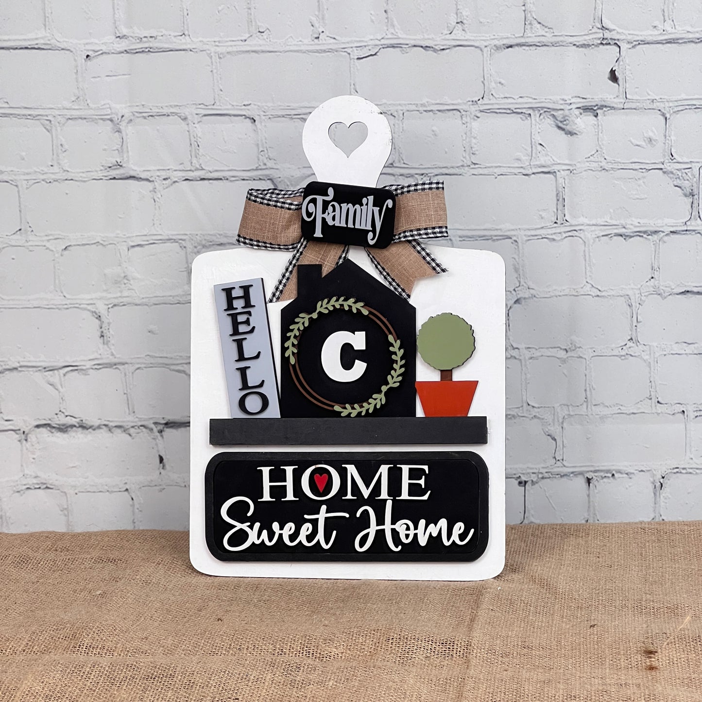 The Breadboard Interchangeable Base with Insert by Janet's Craft Corner features a heart-topped sign with "Family," "Hello," a wreath, a potted plant, and "Home Sweet Home" in bold on burlap against white brick. Ideal for home décor enthusiasts.