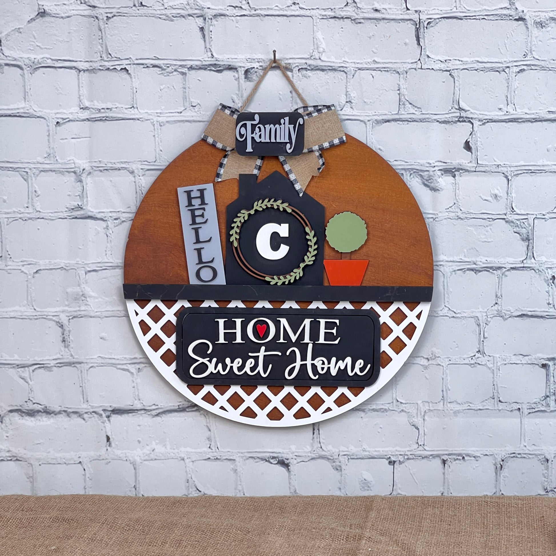 The Janet's Craft Corner DIY kit features a round wooden wall hanging with "Family," "Hello," and "Home Sweet Home" signs, a wreath, letter "C," and orange flowerpot insert. Perfect for brick walls! Kit includes 1 interchangeable door hanger base and 1 insert.