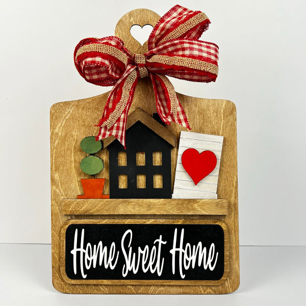 The Breadboard Interchangeable Base with Insert from Janet's Craft Corner is a DIY home decor kit that includes a breadboard-shaped wooden plaque featuring a house, two potted plants, and a heart. It's adorned with a red and white checkered bow and displays "Home Sweet Home" above the chalkboard-style panel.