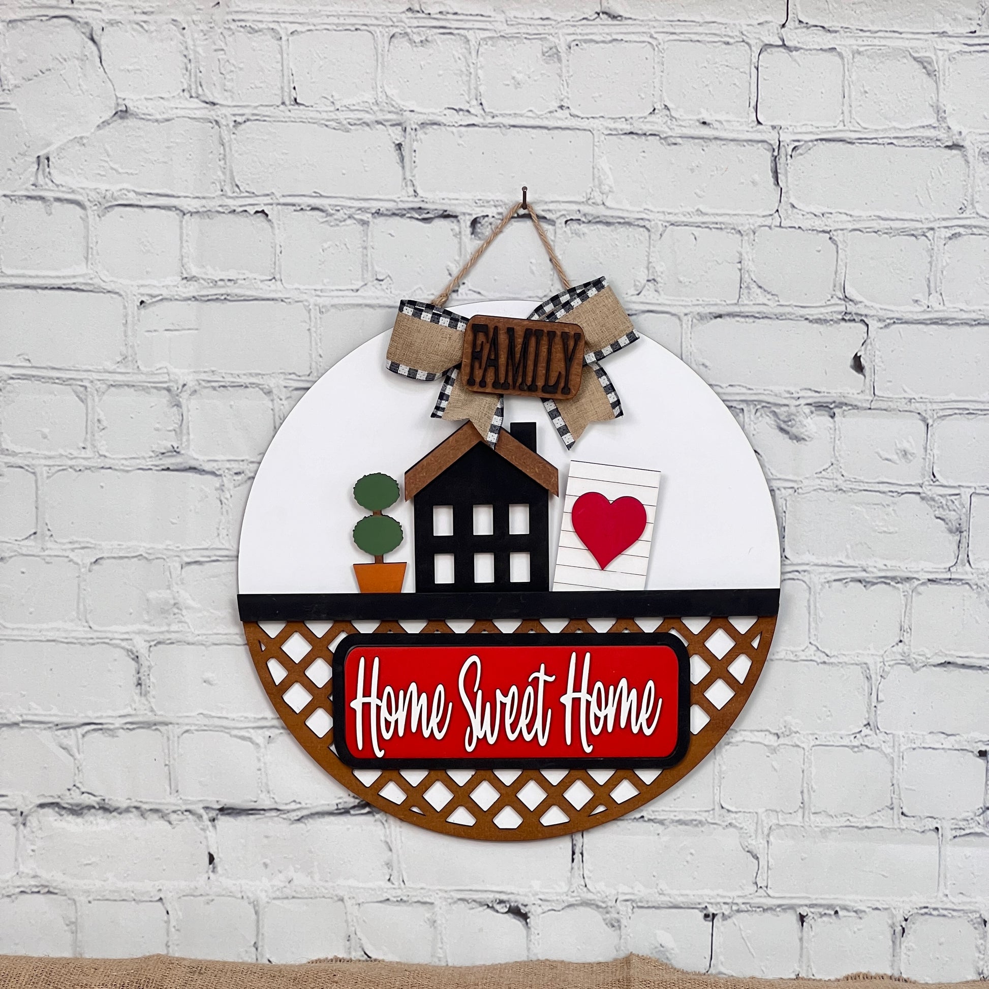 A round decorative sign on a white brick wall showcases a black house, potted plant, heart with a checkered "Family" bow, and "Home Sweet Home" on a red lattice-bordered rectangle. This door hanger is part of Janet's Craft Corner's DIY interchangeable home decor kit collection.