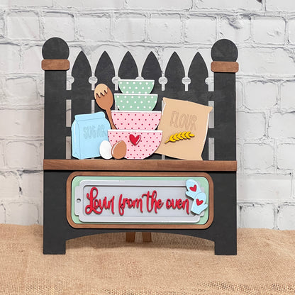 The Garden Fence with Insert from Janet's Craft Corner is a DIY home decor kit featuring an oven-shaped sign with colorful bowls, sugar, flour, eggs, and utensils. The red script reads "Love from the oven" with small hearts. Ideal for craft kit enthusiasts or interchangeable insert lovers.