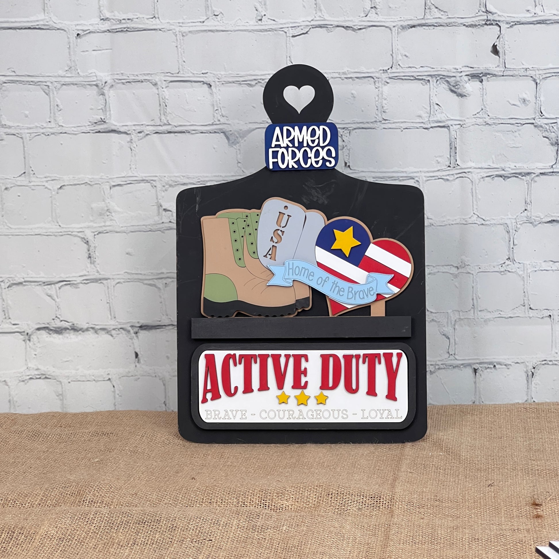 Janet's Craft Corner offers a Breadboard Interchangeable Base with Insert, perfect for DIY home décor. It includes a heart-shaped top labeled "Armed Forces," featuring a boot, USA dog tags, and a star-spangled badge. A sign below reads "Active Duty" with "Brave - Courageous - Loyal.