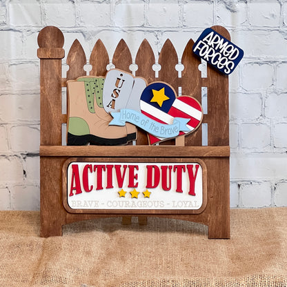 Janet's Craft Corner presents the Garden Fence with Insert, a DIY interchangeable decor kit featuring a picket fence and inserts such as combat boots, a heart with stars and stripes, "USA Home of the Brave," and "ACTIVE DUTY: Brave - Courageous - Loyal." .